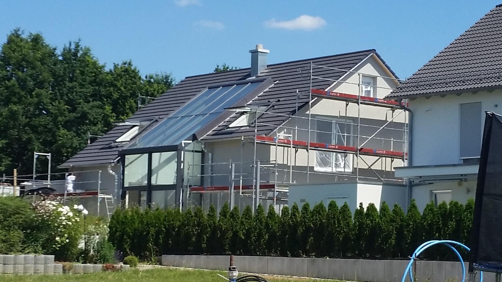 Residential house with scaffolding and solar panels on the roof; garden, hedge, construction workers at work.