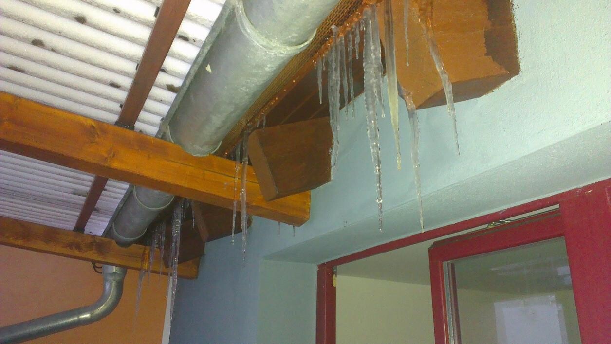 Interior ceiling with wooden beams, piping; icicles hanging next to a red window frame.