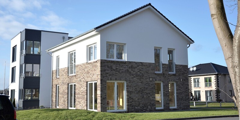 Modern two-storey house with white upper floor, brick base, large windows and garden.