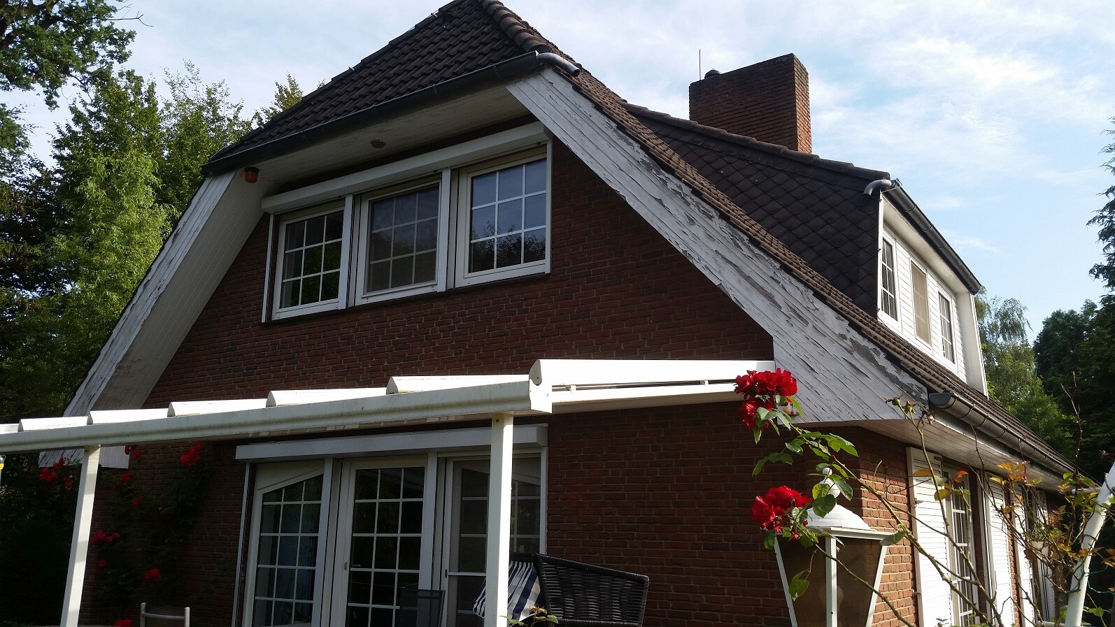 Red brick house with dark roof, white pergola and red roses in the garden.