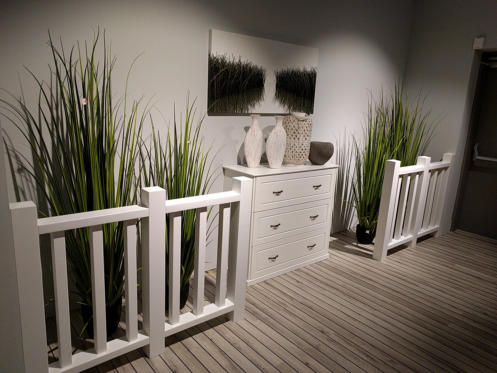 White hallway with chest of drawers, tall grass plants, and wooden plank flooring.
