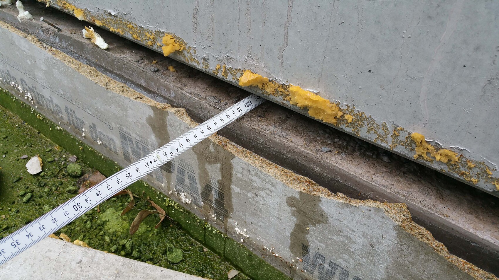 Concrete foundation with damaged edge, yellow sealing compound and measuring tape visible, mossy ground