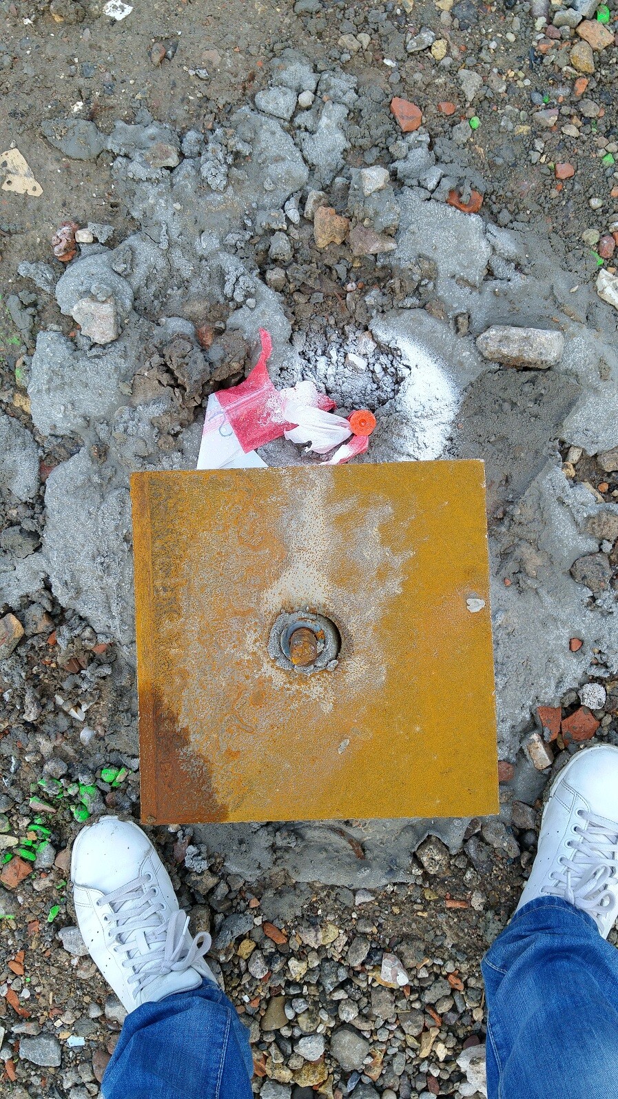 Baustelle mit Schutt, rostigem Metalldeckel und quadratischem Metallteil, Schuhe sichtbar
