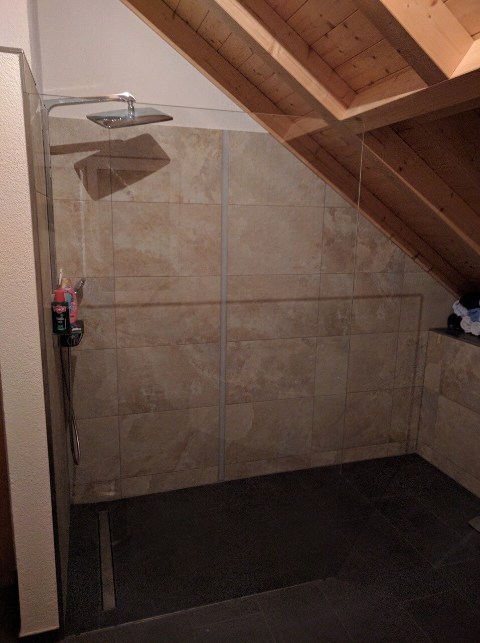 Shower area with glass partition, beige stone tiles, dark floor, wooden roof truss.