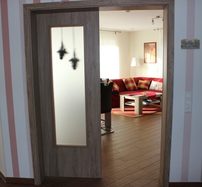 Open wooden door with glass panel leads to modern living room with red sofa.