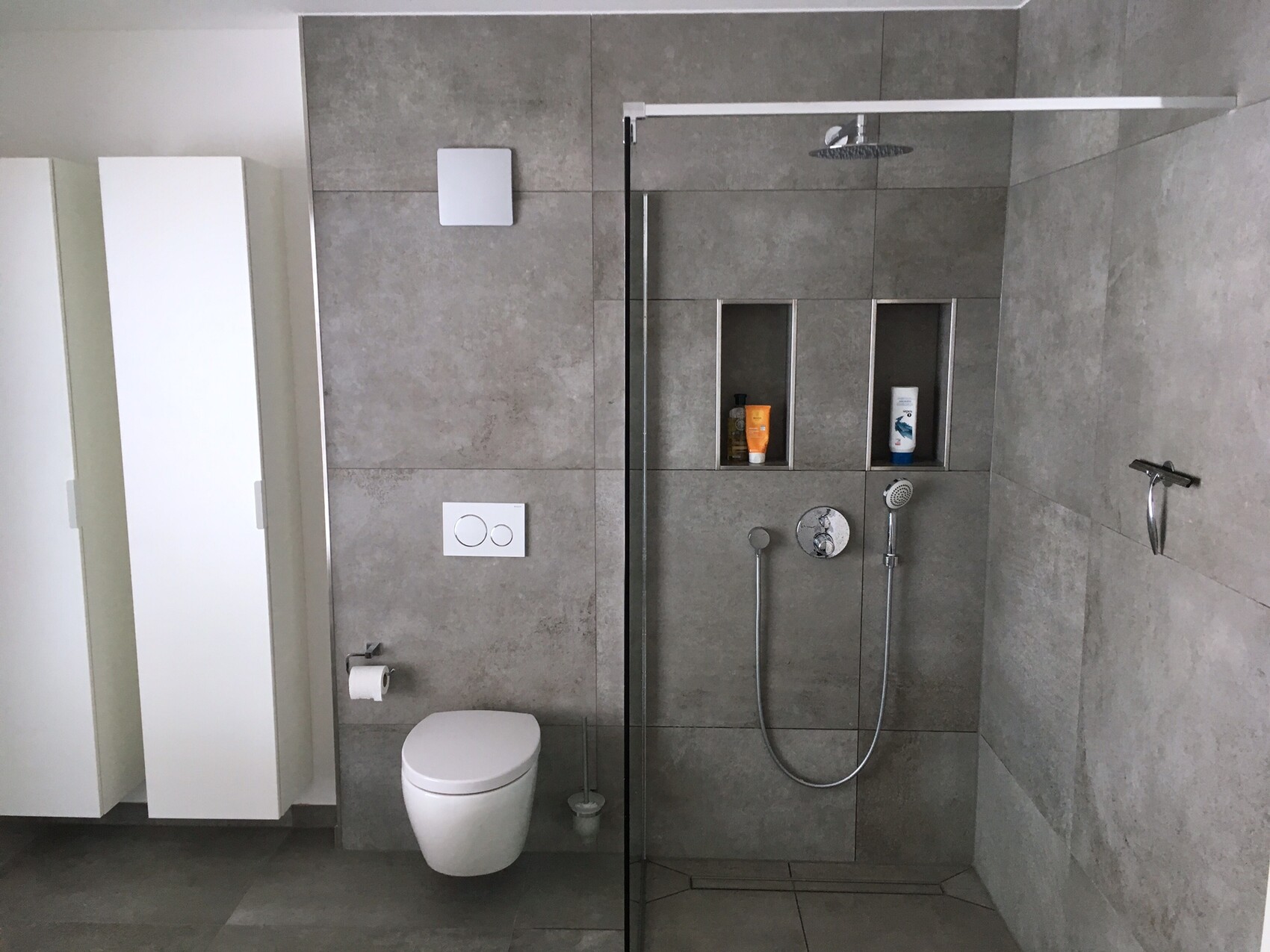 Modern bathroom with gray tiles, wall-hung toilet, glass shower, and shelves.
