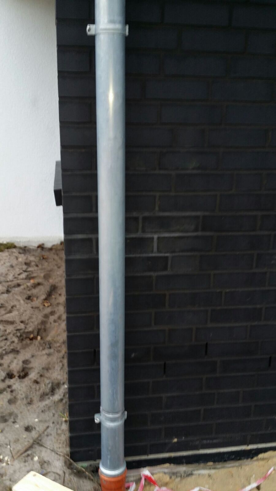 Metal rainwater pipe mounted on a dark brick wall of a house from outside.