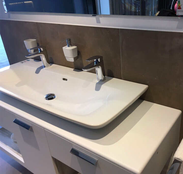 White bathroom vanity with double faucet and soap dispenser in front of grey tile wall.