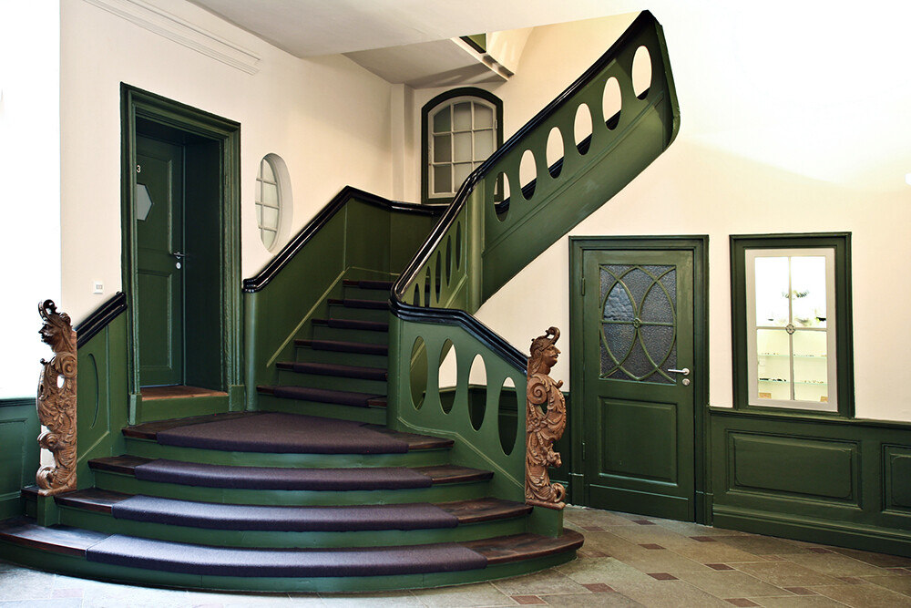 Grüne Wendeltreppe mit dunklem Geländer, geschnitzte Pfosten, grüne Türen im Innenraum.