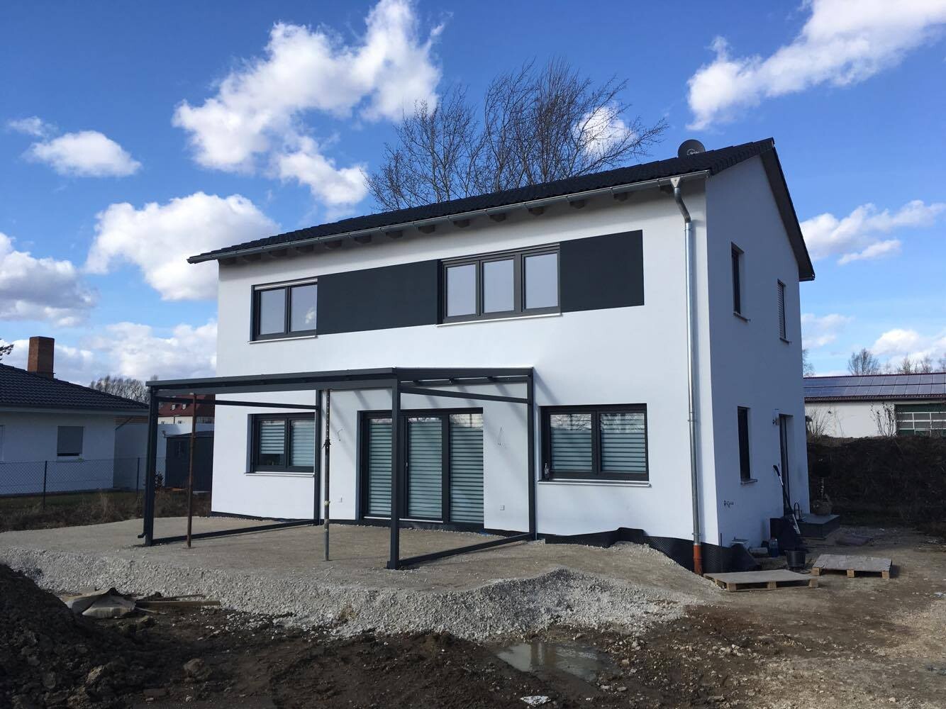 Weißes zweistöckiges Neubauhaus mit dunklen Fensterbändern und Metallcarport; Baustelle.