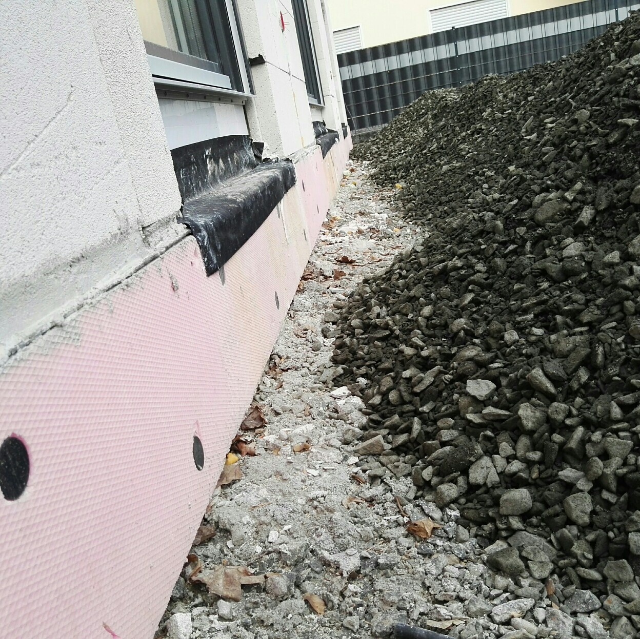 Außenwand eines Baugrundstücks: pinke Dämmplatten an der Fassade, Fenster links, Kieshaufen rechts.