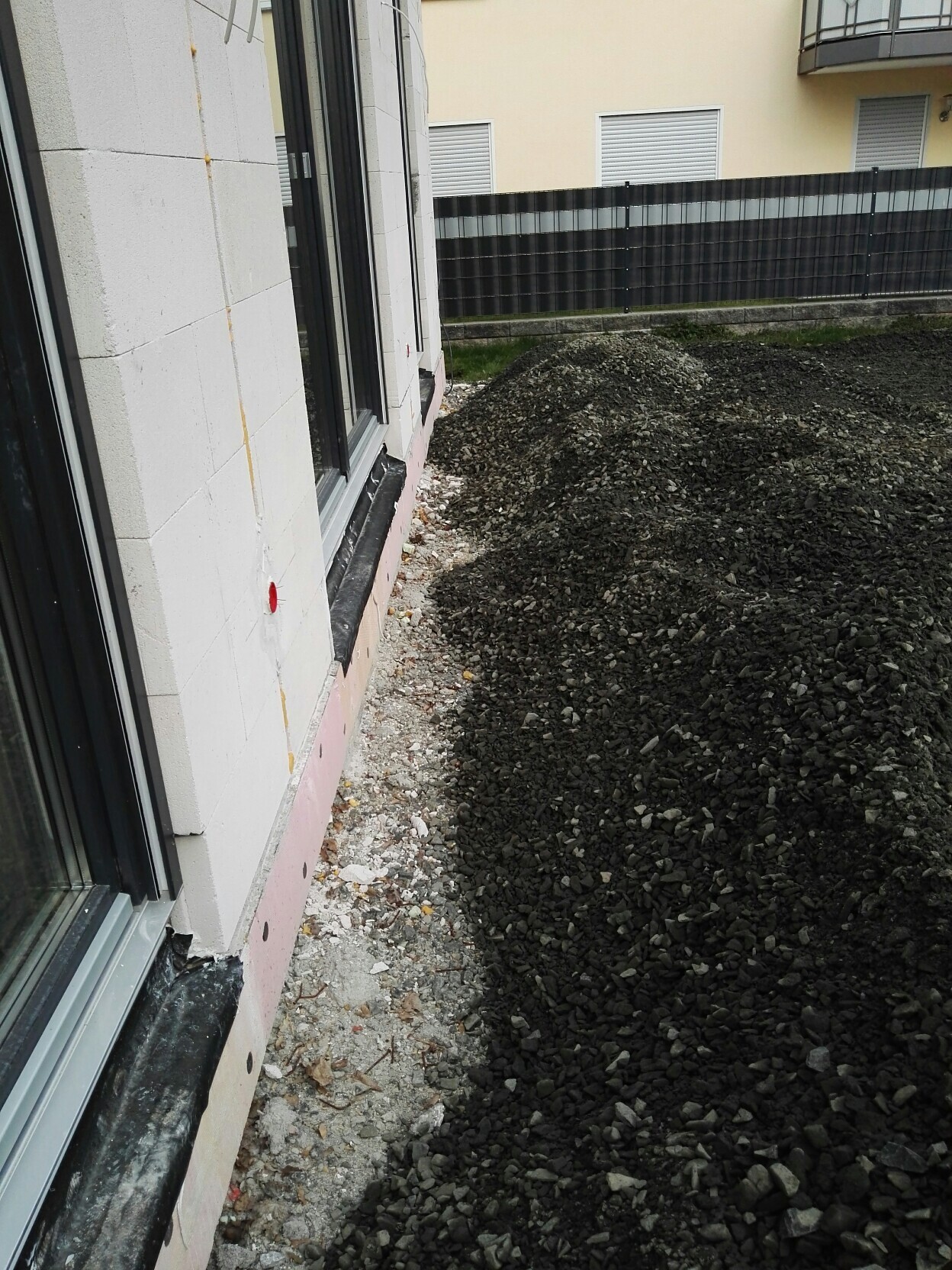 Außenansicht eines Gebäudes; Fensterrahmen sichtbar, großer Kieshaufen neben der Wand.