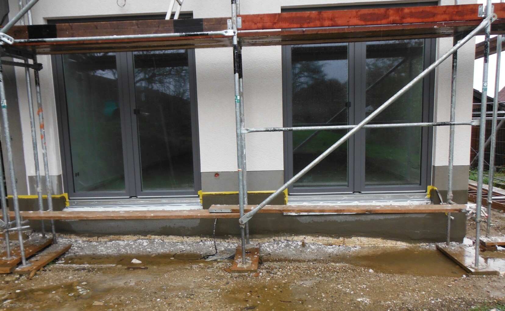 Baustelle vor Haus: Große Glasschiebetüren hinter Stahlgerüst, schlammiger Boden.