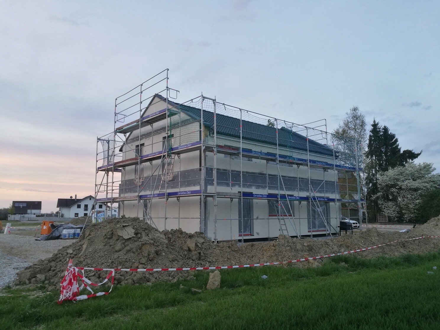 House in shell construction surrounded by scaffolding; piles of earth and barrier tape in front of the construction site.