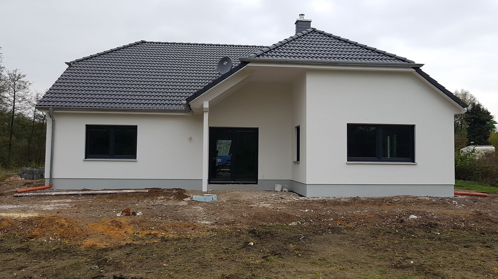 White single-family house with dark tiled roof, construction work on the future site