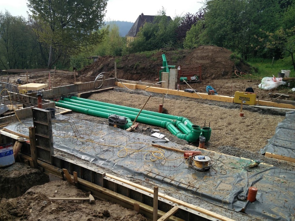 Construction pit with foundation work, green pipes laid out, and construction tools visible.