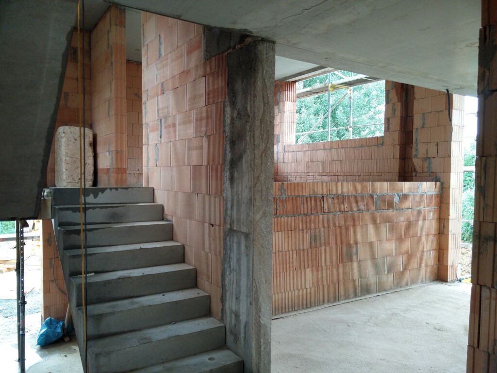 Baustelle: Innenraum mit rotem Ziegelmauerwerk, Betonstufen und Stütze, offenes Fenster.