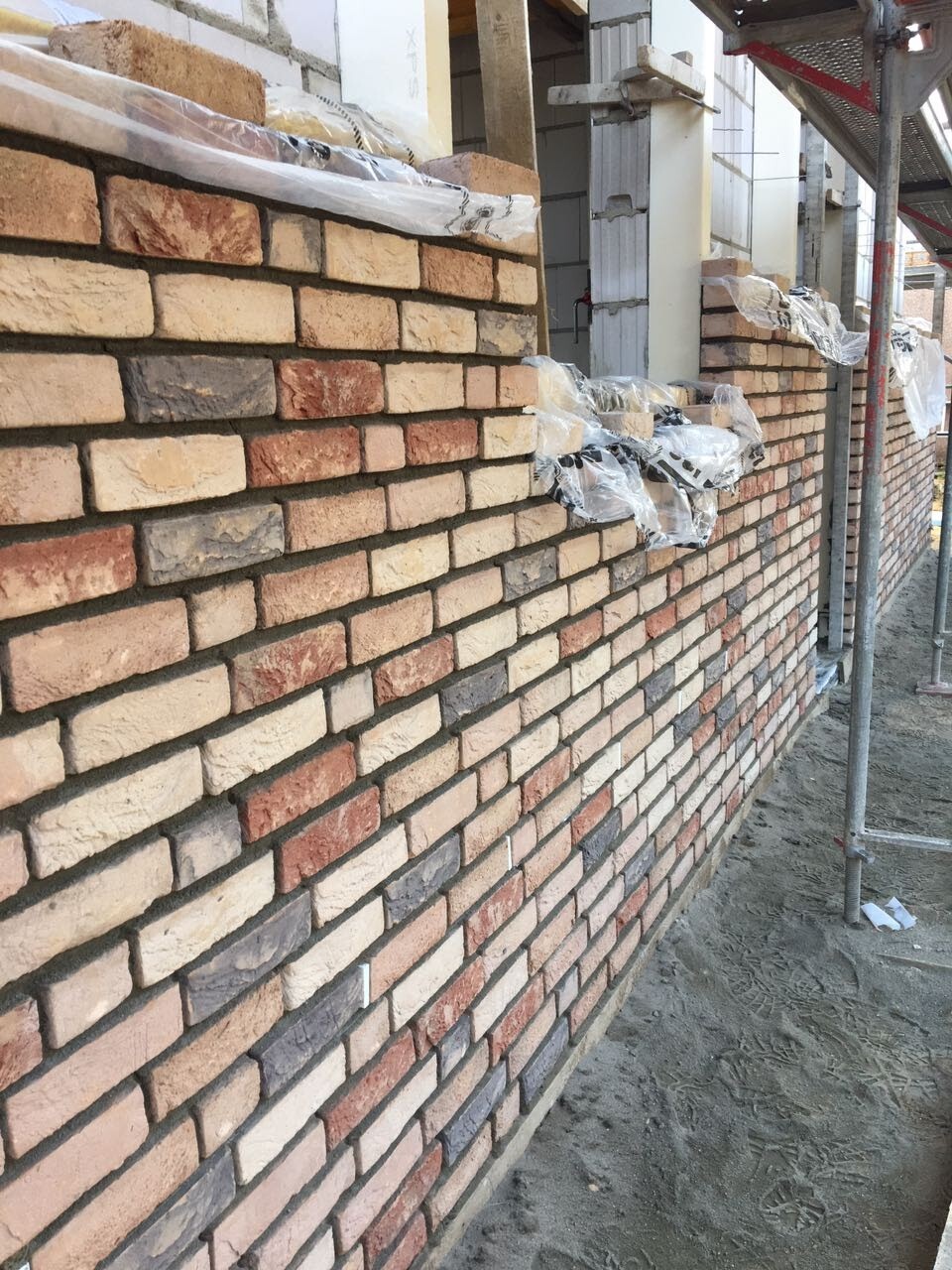 Brick wall under construction with scaffold poles and bricks stacked in plastic wrap.