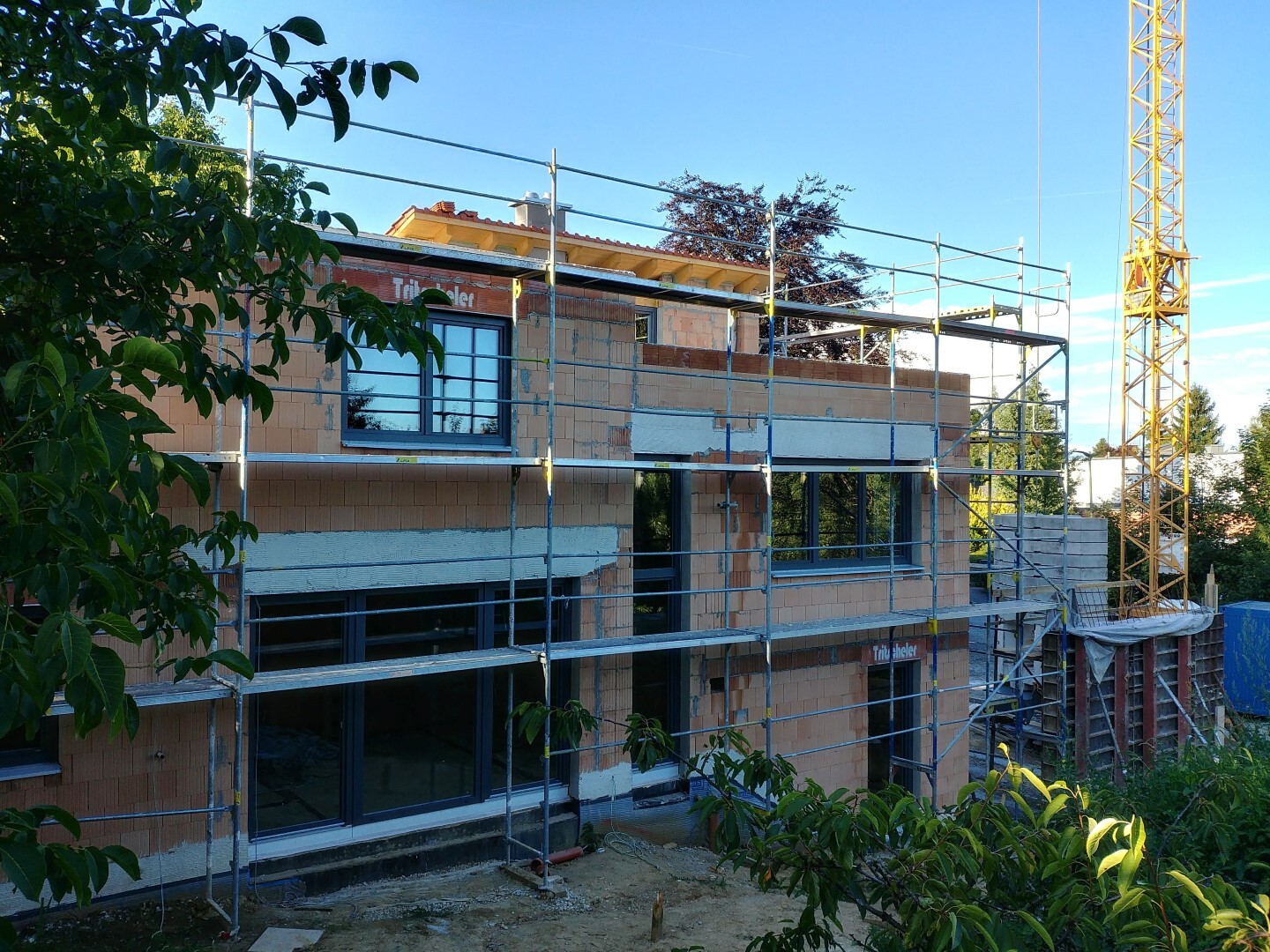 Multi-story building shell with scaffolding, construction crane on the right, green trees in the foreground