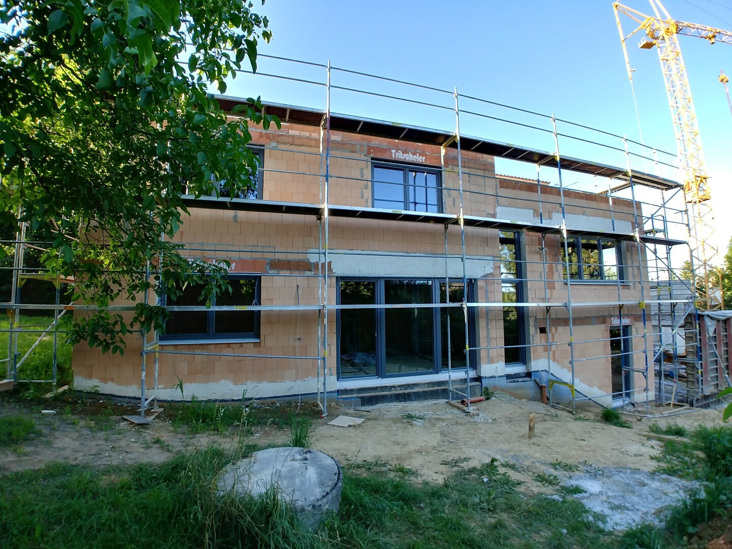 Two-story building shell with scaffolding, crane on the right, tree on the left.