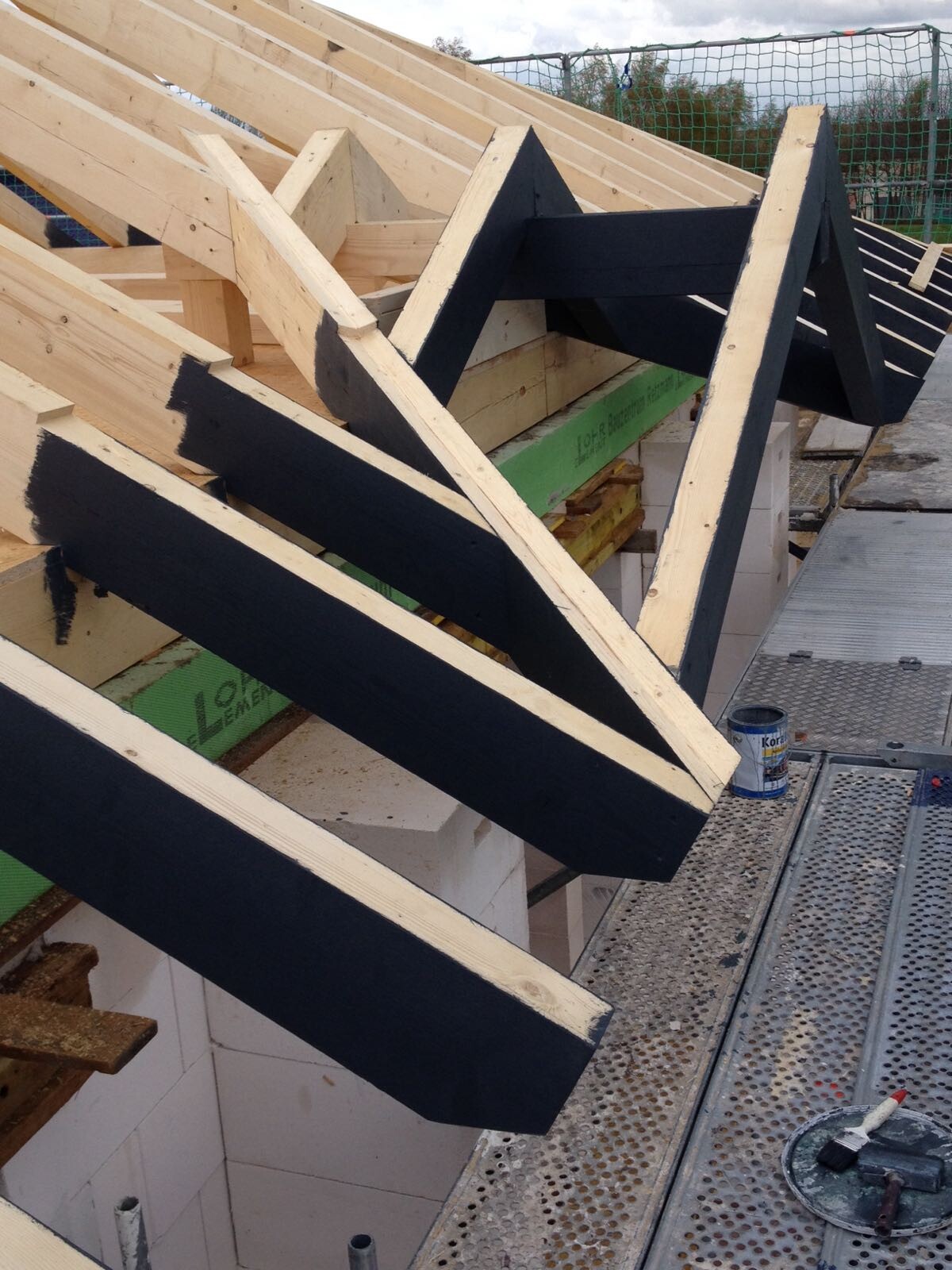 Roof structure made of angled wooden beams, painted black, on scaffolding.