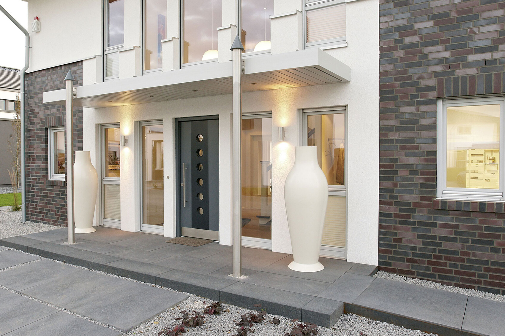 Modern house facade with dark door, glass windows, and two large white designer vases at the entrance.
