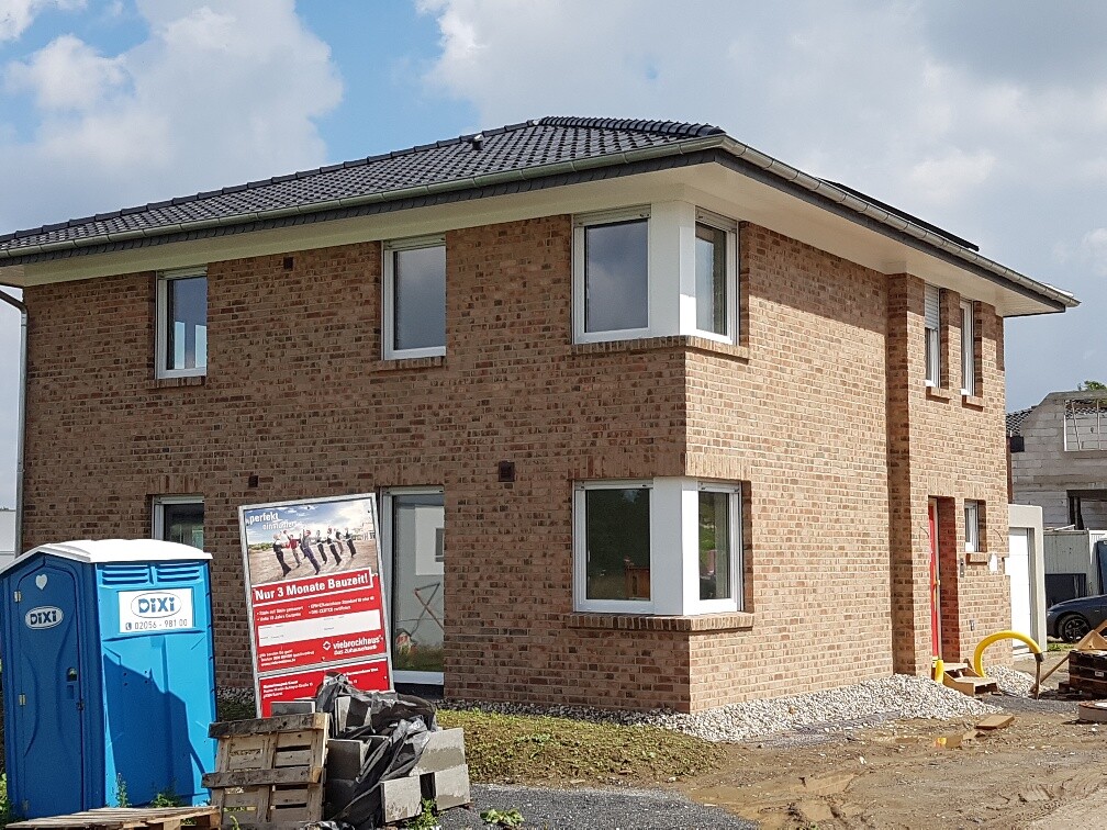 Two-story brick house under construction with construction equipment, blue portable toilets, and building materials.