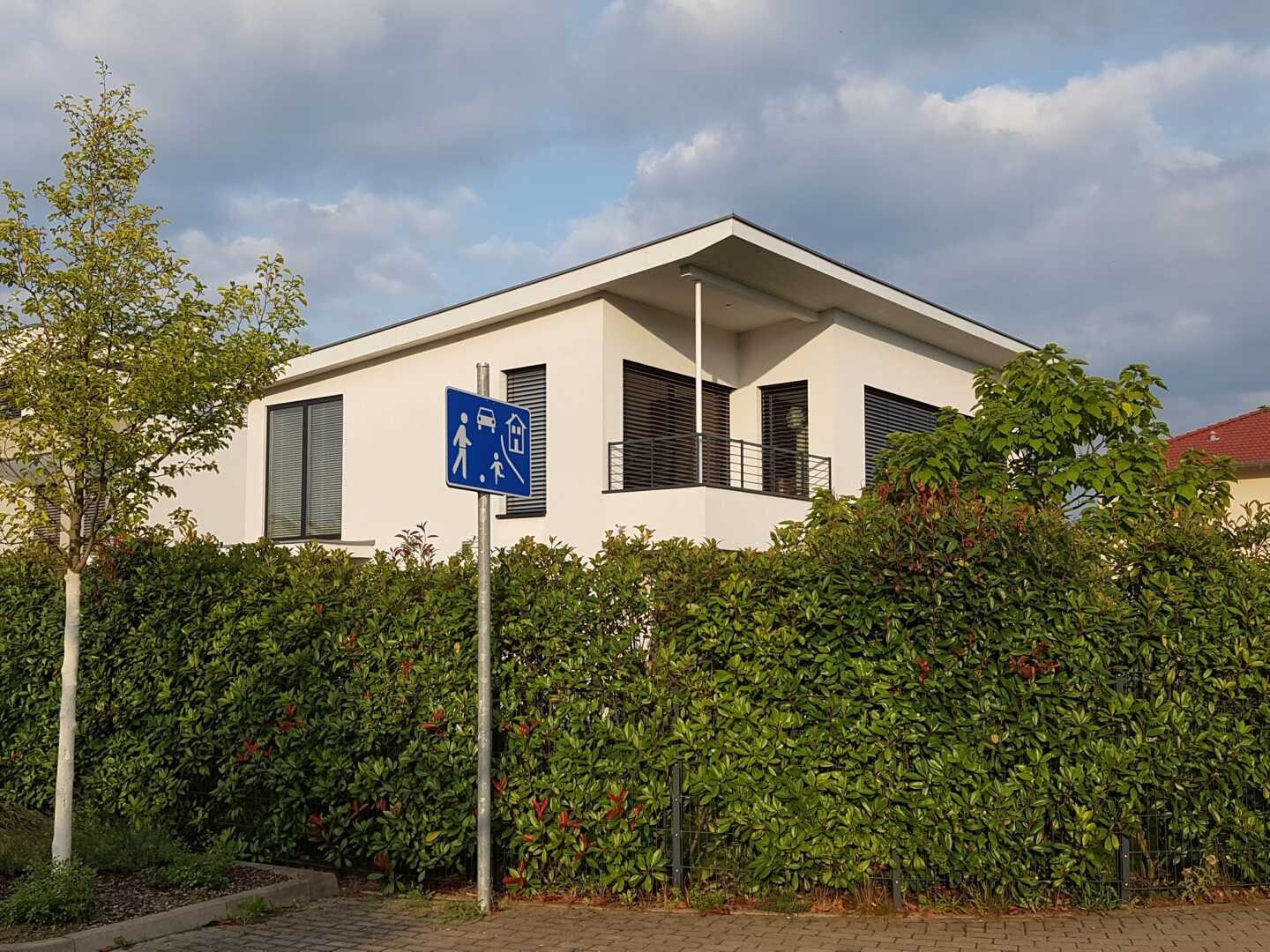 Modern white house behind hedge; balcony visible, blue pedestrian sign at road side, cloudy