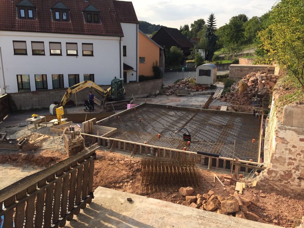 Construction site with foundations, reinforcement, excavator, construction workers, and surrounding houses.