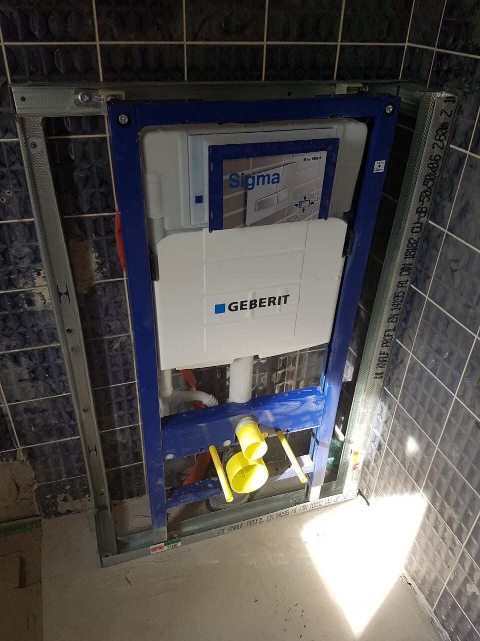 Geberit Sigma concealed cistern in metal frame of pre-wall installation, yellow pipe connections visible.