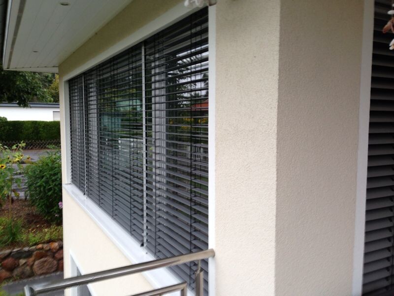 Exterior window of a house with horizontal blinds, beige wall and garden.