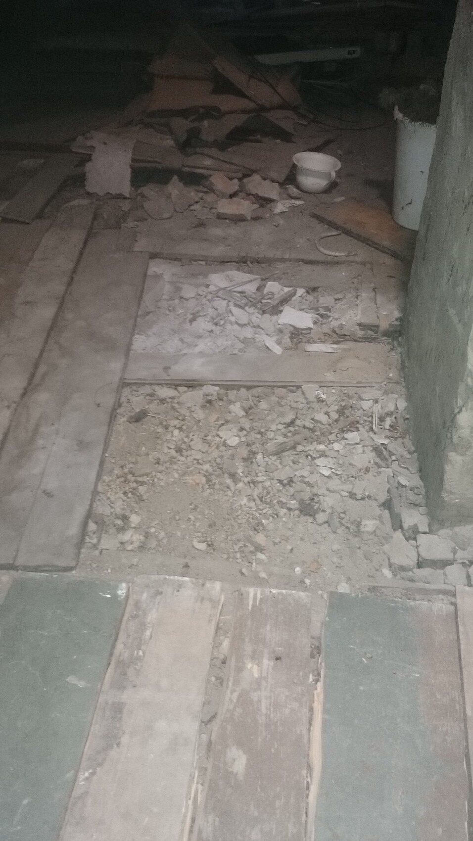 Dunkler Raum mit bröckelndem Boden aus Holzbrettern, Staub und Geröll; eine weiße Tasse liegt daneben