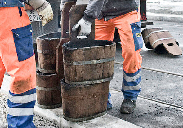 Zwei Arbeiter in orangefarbener Schutzkleidung heben Holzfässer auf der Straße neben Gleisen.