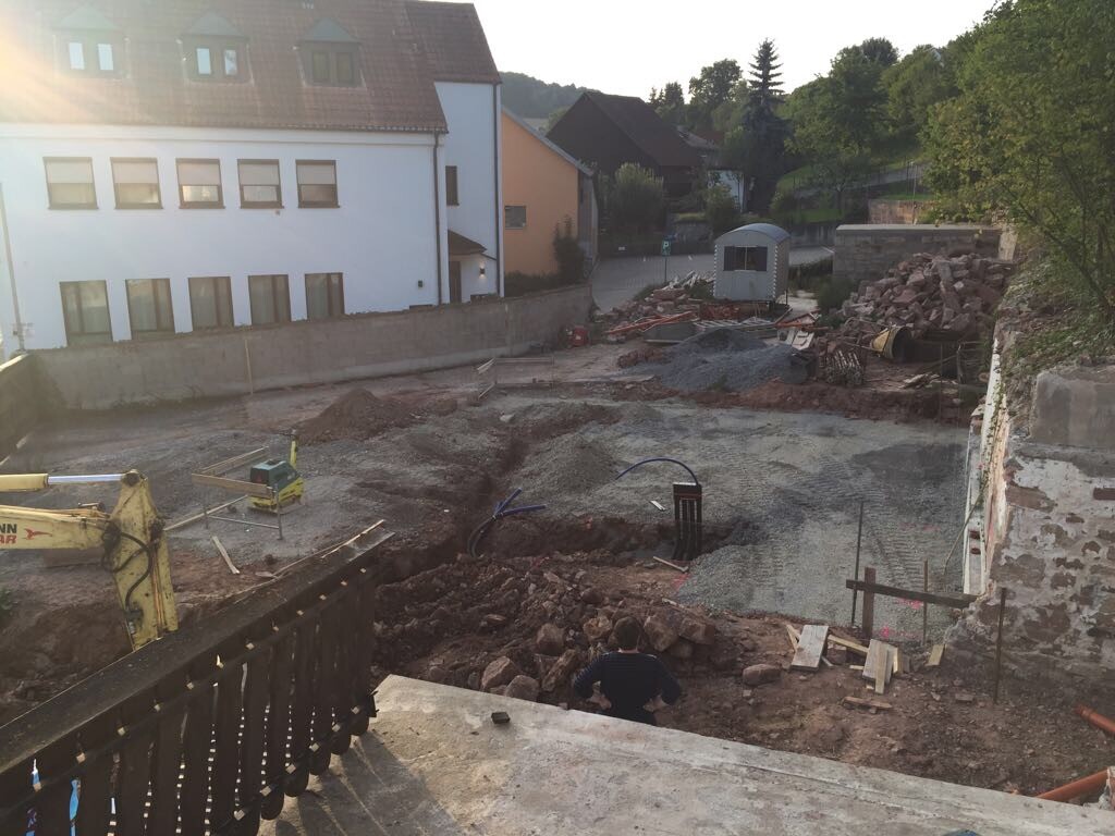 Construction site: excavation pit, gravel, excavator, building materials; row of houses in the background.