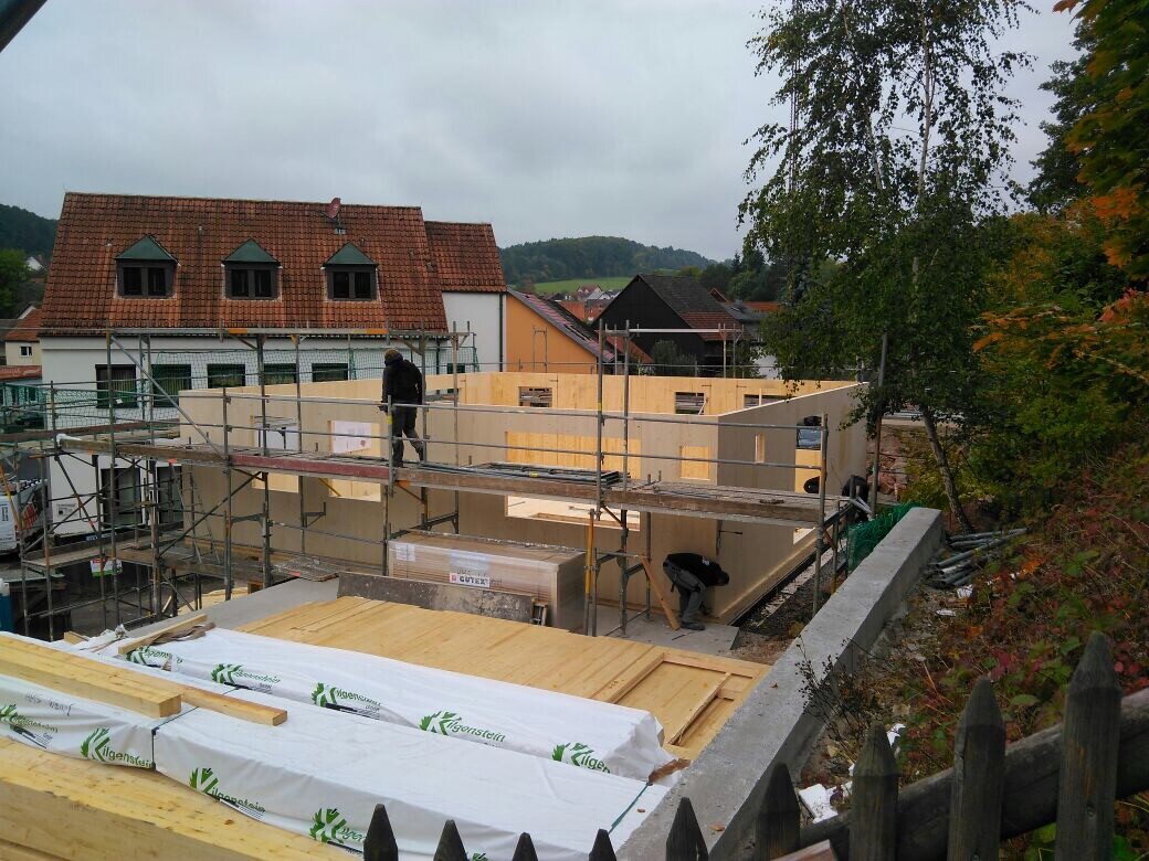 Construction site with wooden walls, scaffolding, and worker; surrounding houses and trees.
