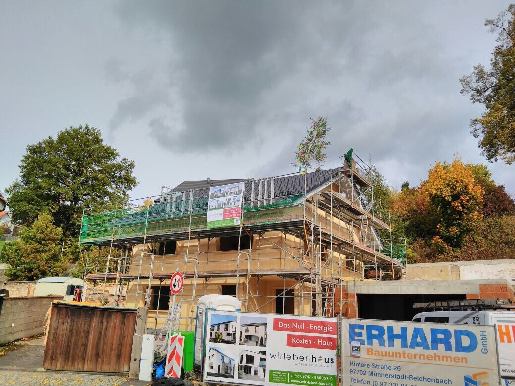Two-story house in shell construction with scaffolding, construction fence, and banner at the site.