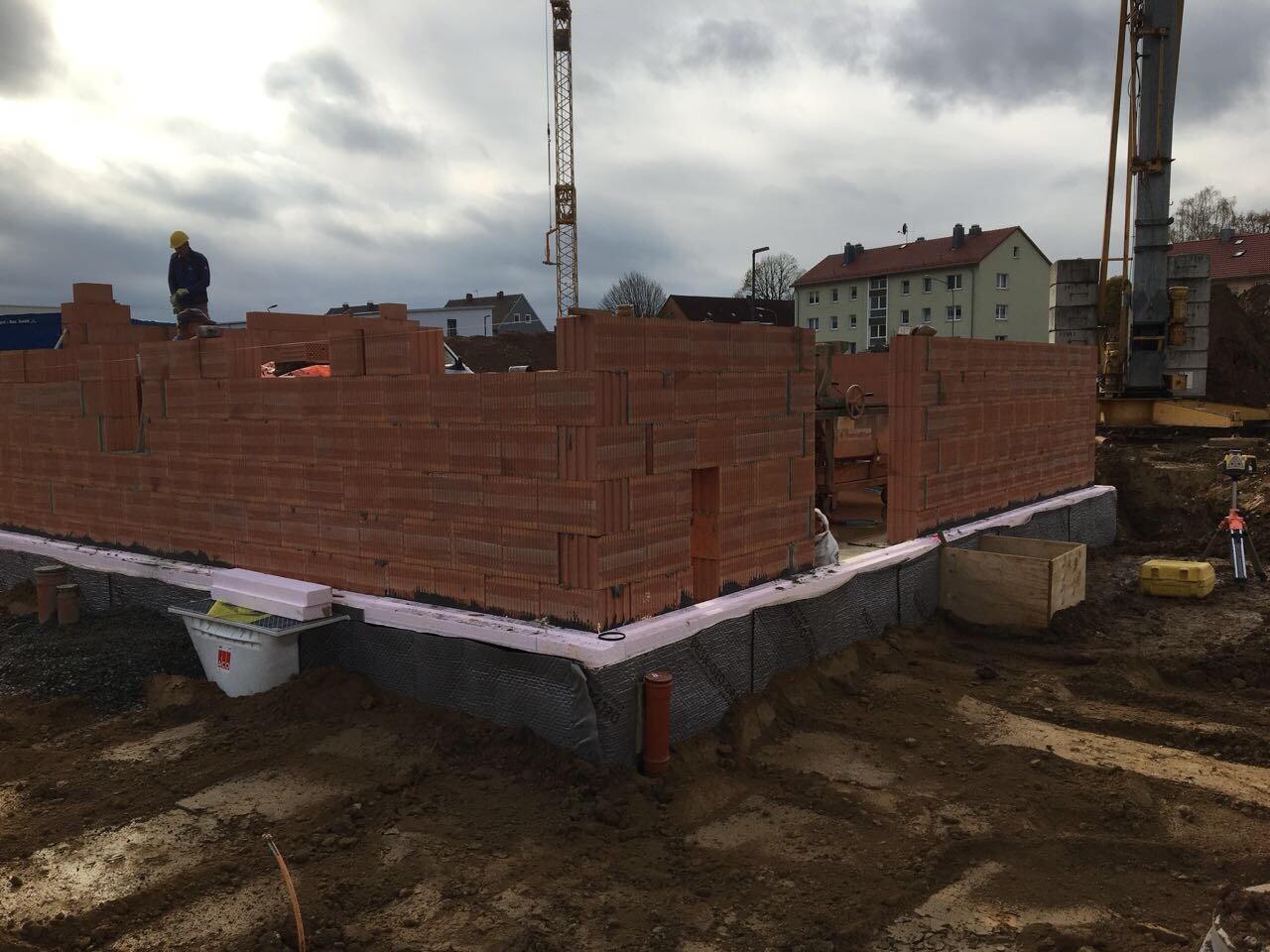 Building site with red brick walls on foundation; construction workers with helmets and crane in the background.