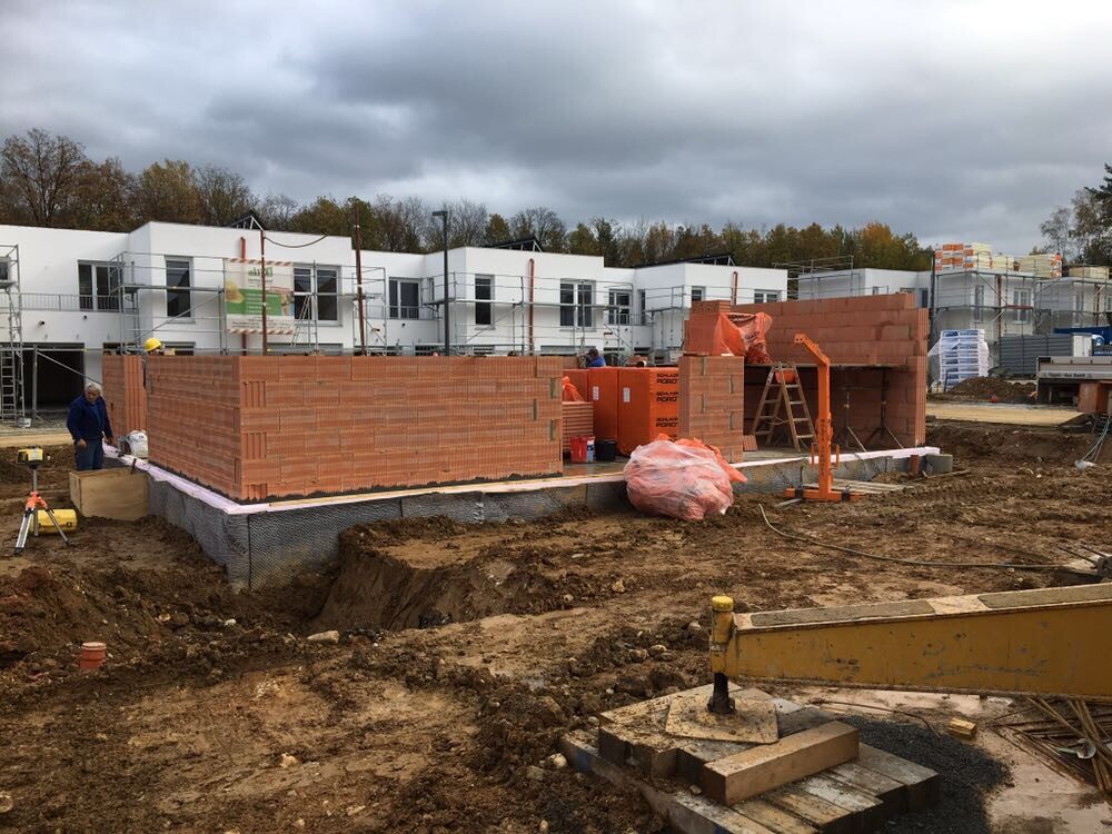 Construction site with red brick building, construction worker, scaffolding, soil and white buildings in the background.