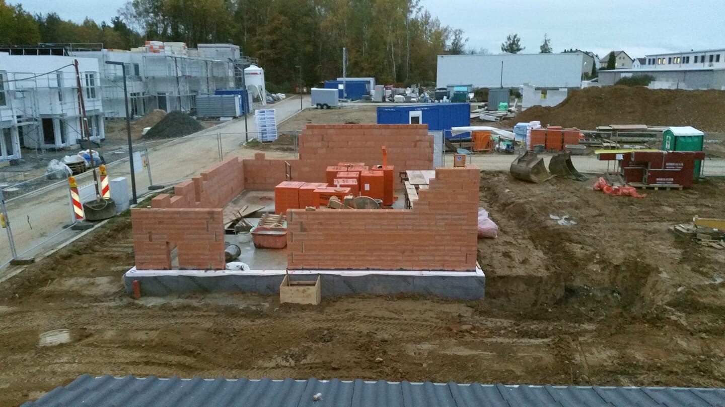 Shell of a small house made of red bricks on a construction site, earthworks visible.