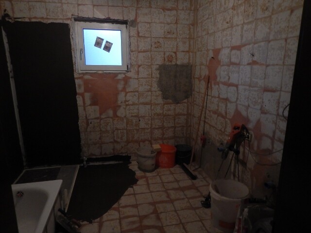 Unrenovated bathroom with tile remains, buckets, tools, and a small window.