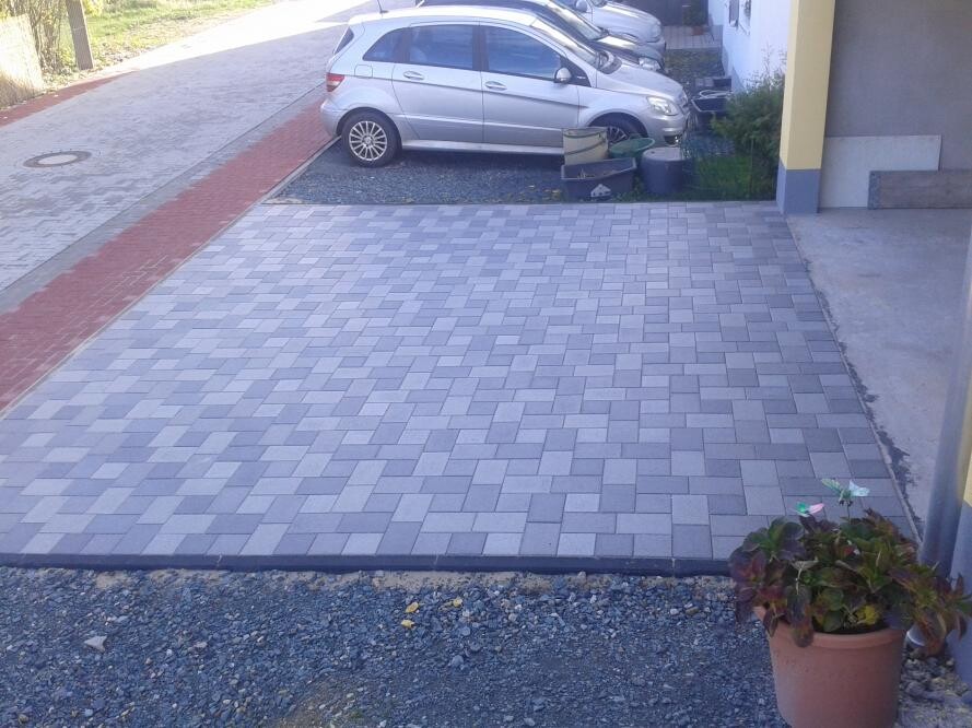 Large gray paved area in the foreground; parked silver car behind, potted plant on the right.