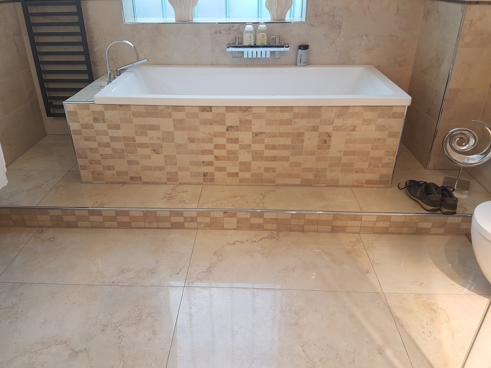 Large bathtub with beige mosaic tiles, window behind; pair of shoes next to spiral-shaped stool.