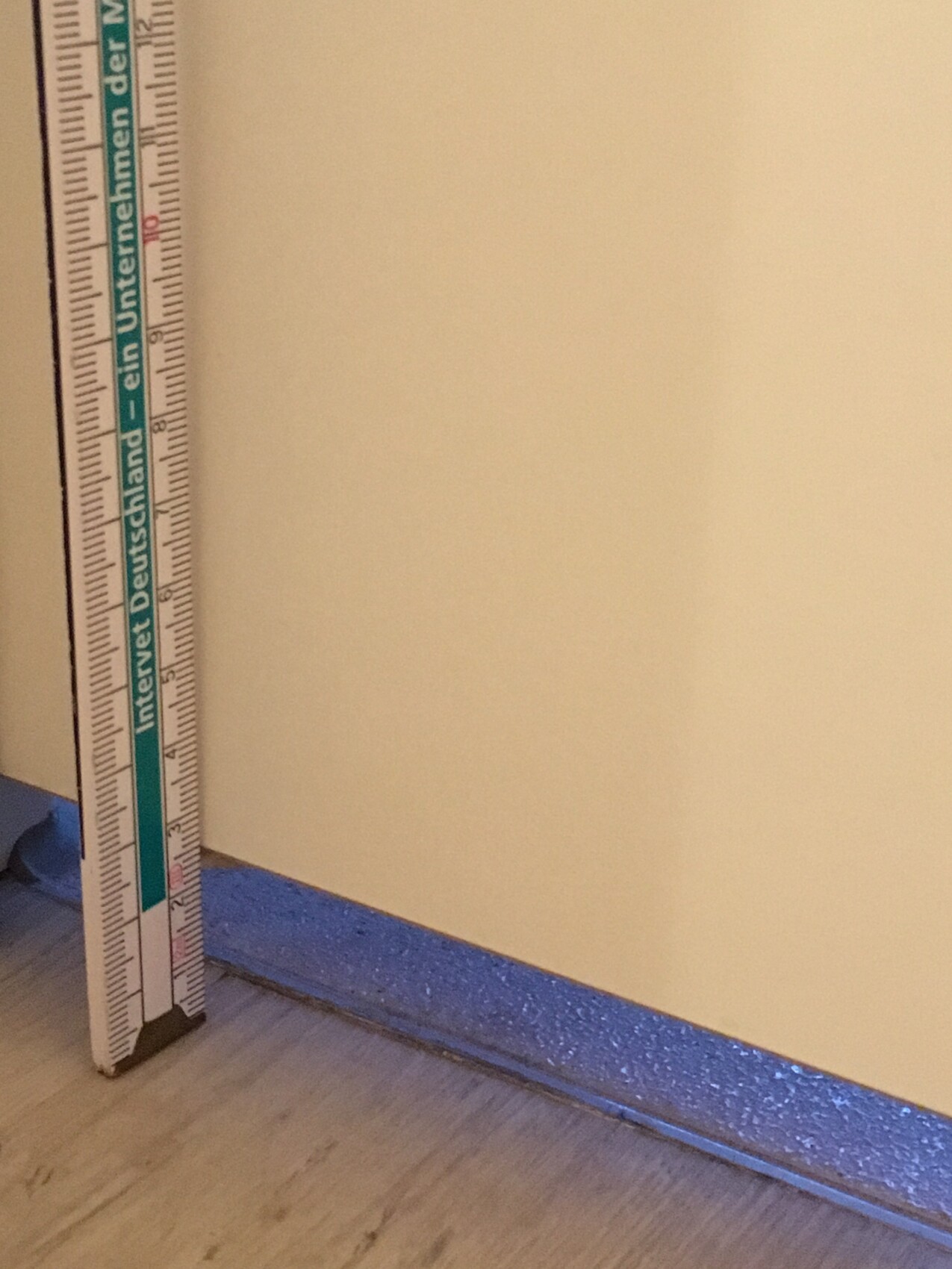 Messlatte steht senkrecht an heller Innenwand neben blauem Sockel im Innenraum.