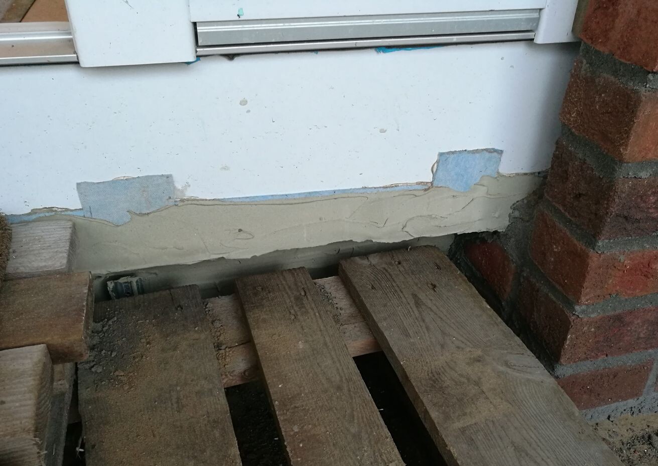 Exterior wall with flaking plaster and wooden pallets in front, visible foundation.