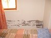 Small bedroom with bed, window and orange curtains, wall partly damaged.