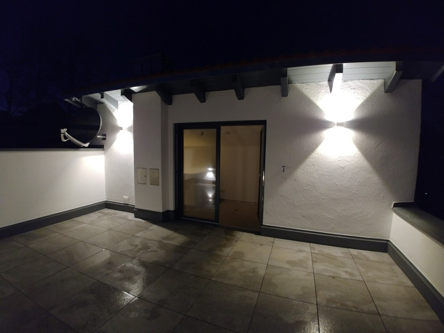Night view of a roof terrace with glass sliding door, wall lamp, and satellite dish.