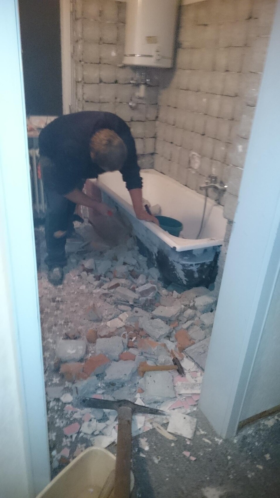 Worker removing tiles in the bathroom, construction site with debris and bathtub visible.
