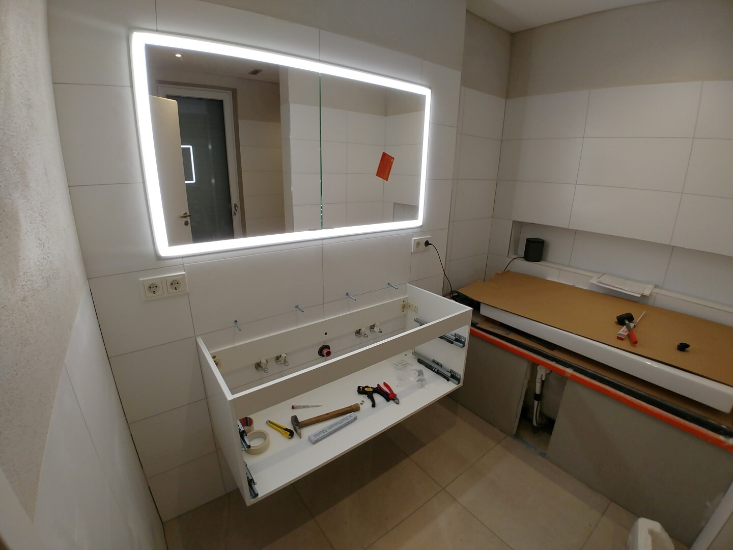 Modern bathroom under renovation: illuminated mirror, open washbasin with tools, bathtub.