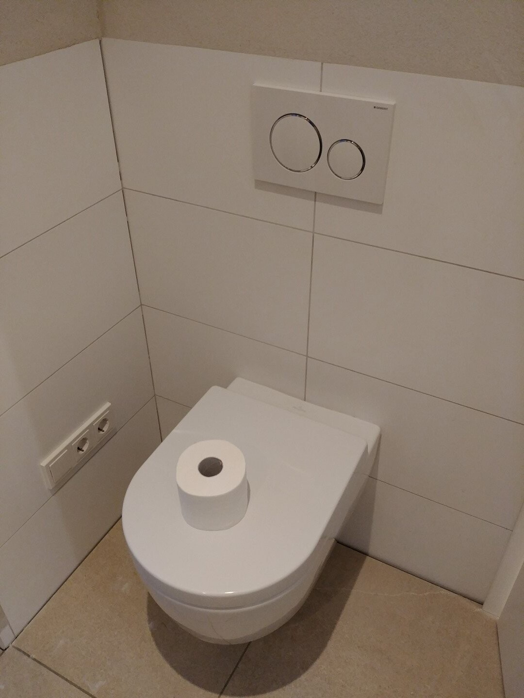 Toilet with wall-mounted dual-flush cistern, lid with toilet paper roll, white tiles.