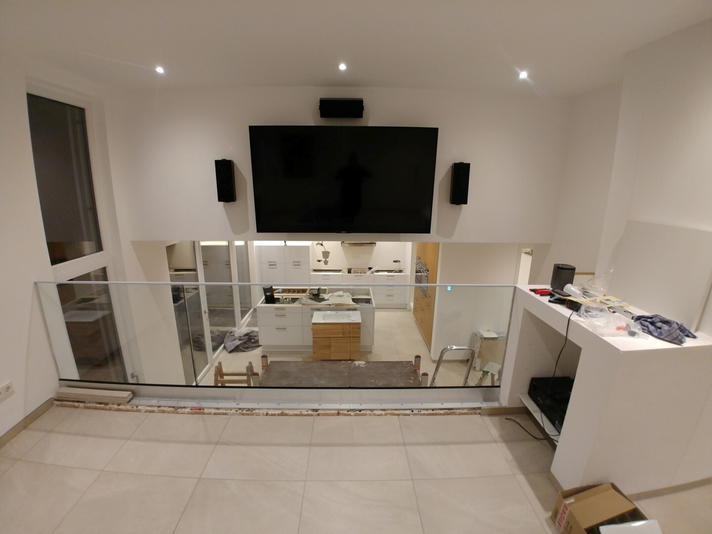 Large flat-screen TV on white wall, speakers on the side; glass railing over the kitchen area.